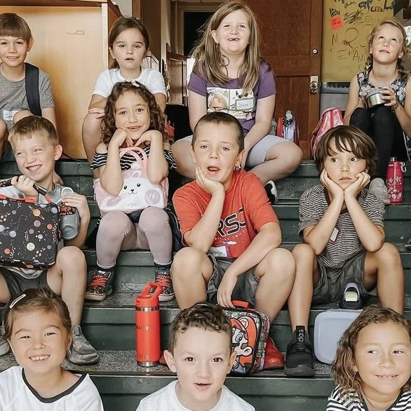 Children sitting together on steps with lunch bags and water bottles