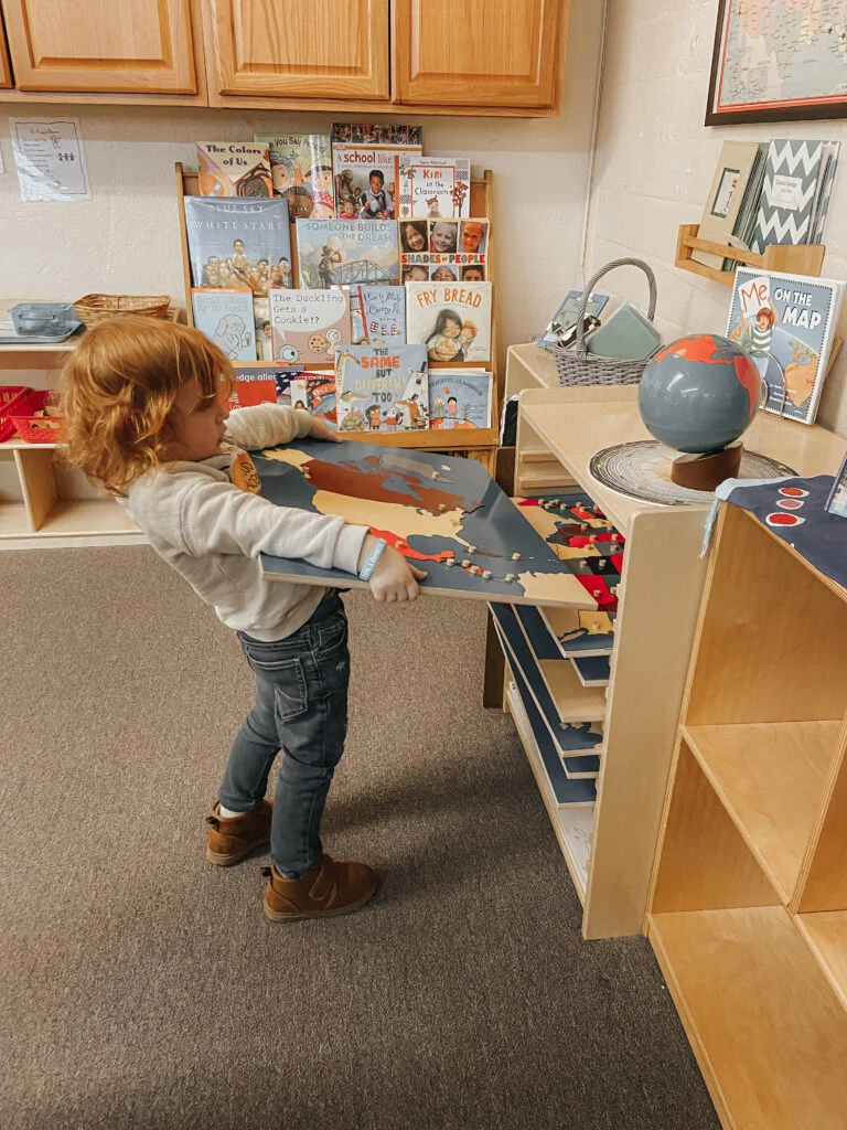 Child carrying a map puzzle beside a globe and shelves of maps