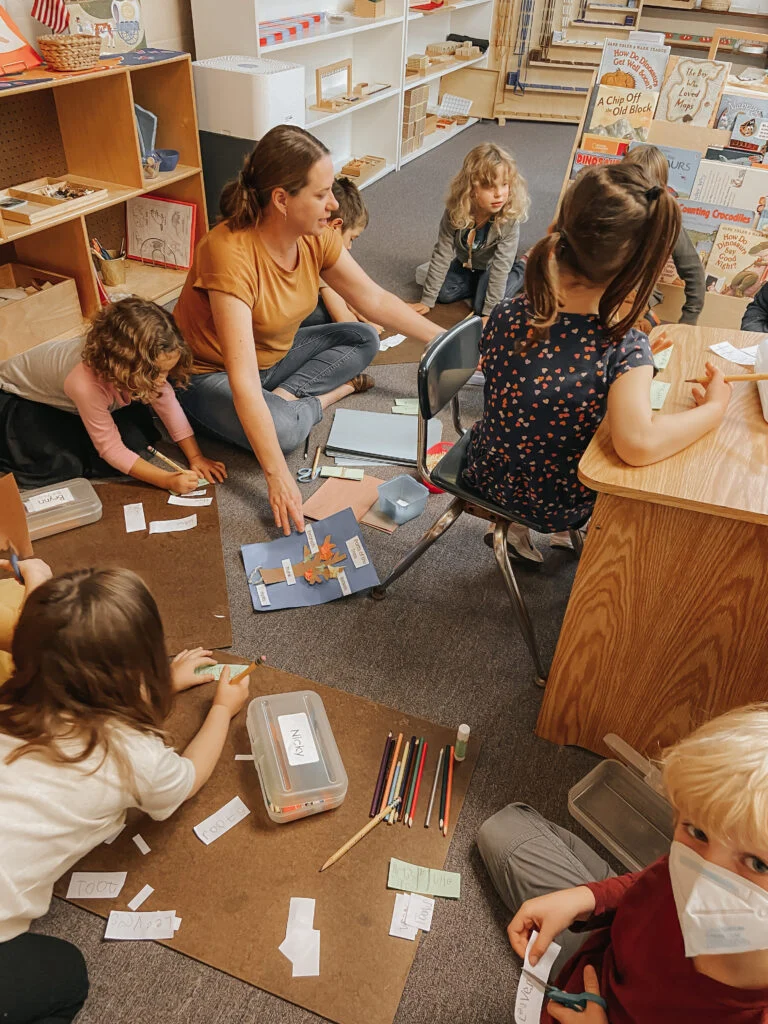 Children writing on paper labels beside an adult seated on the classroom floor