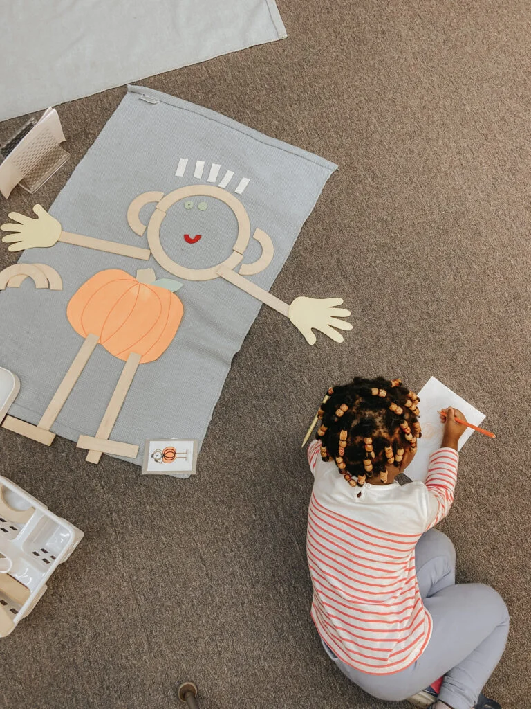 Child drawing beside a figure assembled from shapes on a floor mat