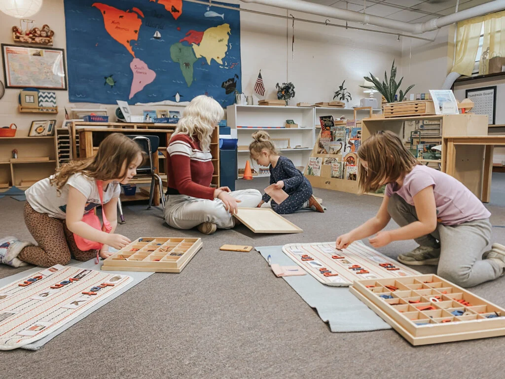 Children arranging movable letters on floor mats beside a seated adult
